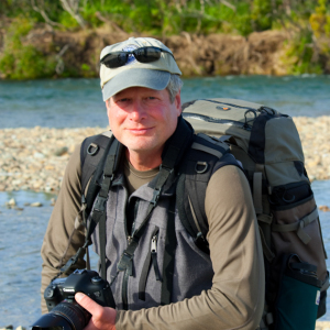 Man in hiking gear holding camera and wearing a baseball cap